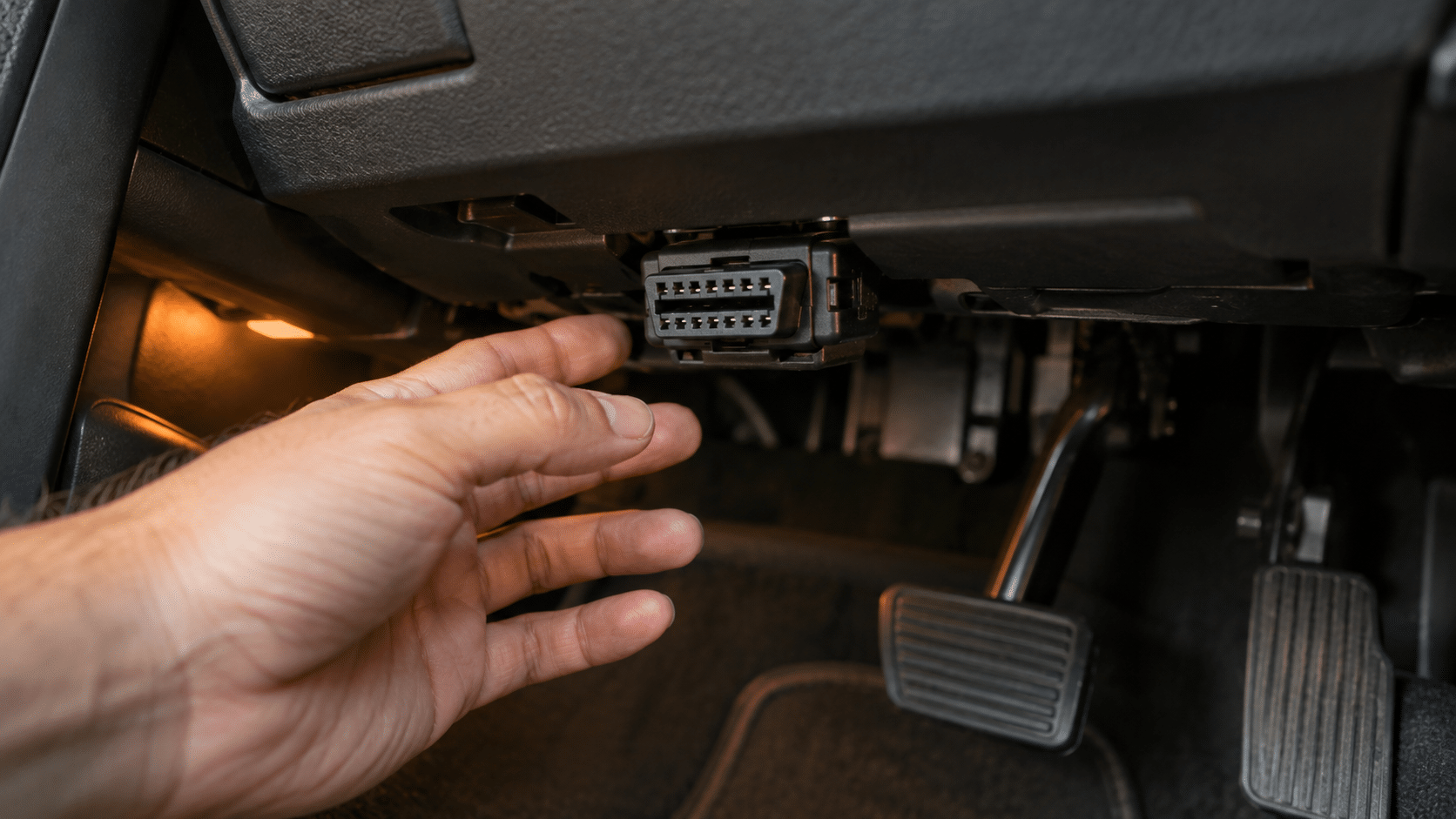 Close-up of a hand reaching toward the 16-pin OBD2 port under the driver-side dashboard.